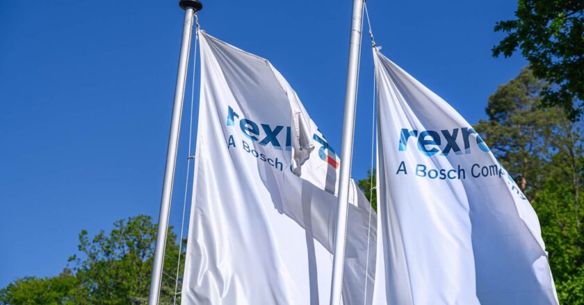 Zwei weiße Flaggen mit dem "Rexroth - A Bosch Company" Logo wehen im Wind vor einem klaren blauen Himmel und grünen Bäumen.