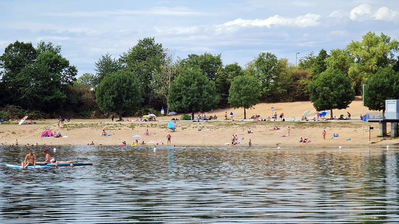 Besucher genießen einen sonnigen Tag am Rotter See in Troisdorf, mit Blick auf das sandige Ufer, umgeben von grünen Bäumen und Leuten, die sich beim Schwimmen und Sonnenbaden entspannen.