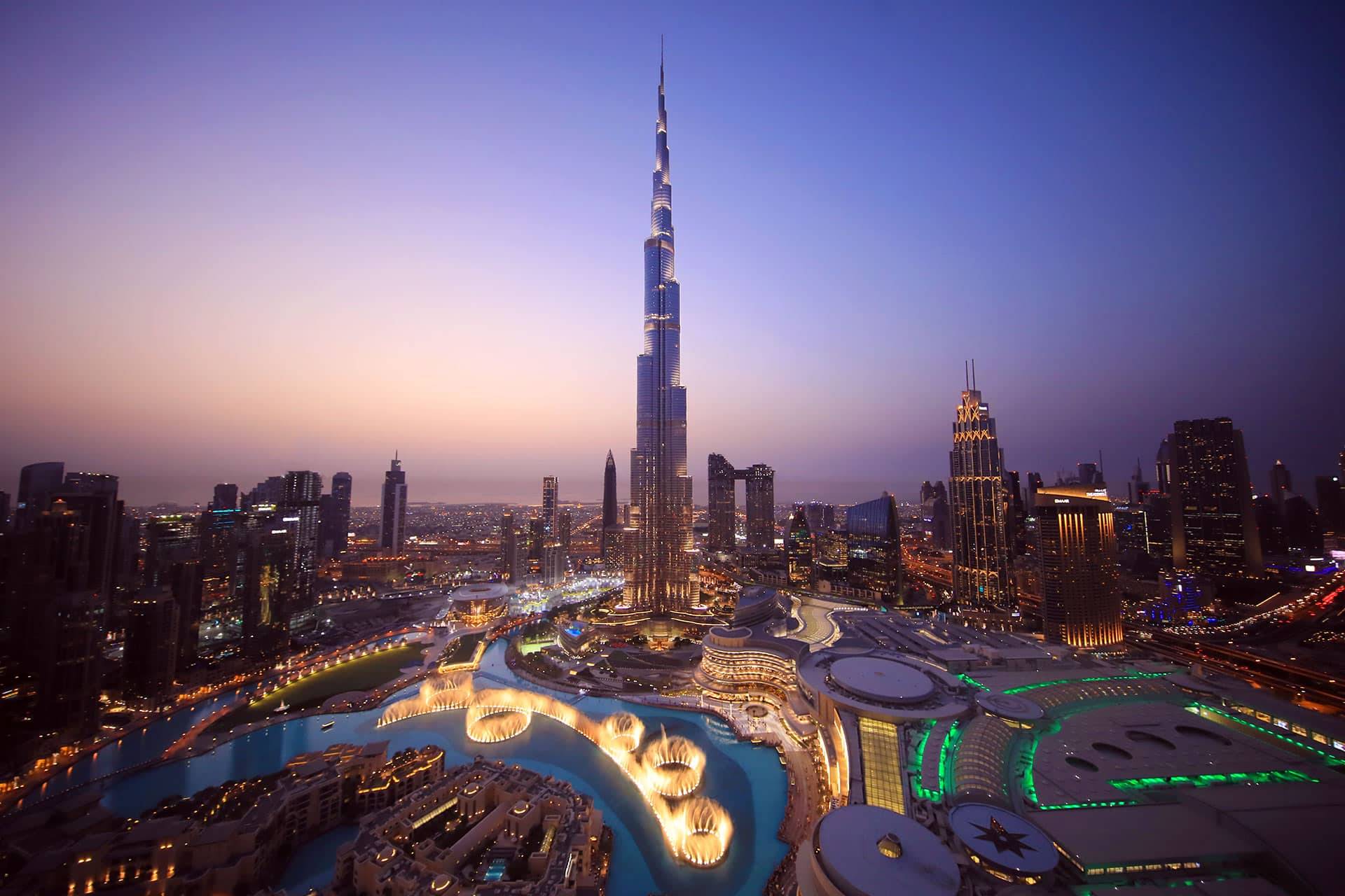 Das Foto zeigt den Burj Khalifa in Dubai bei Dämmerung, umgeben von der beleuchteten Skyline und den Wasserspielen.