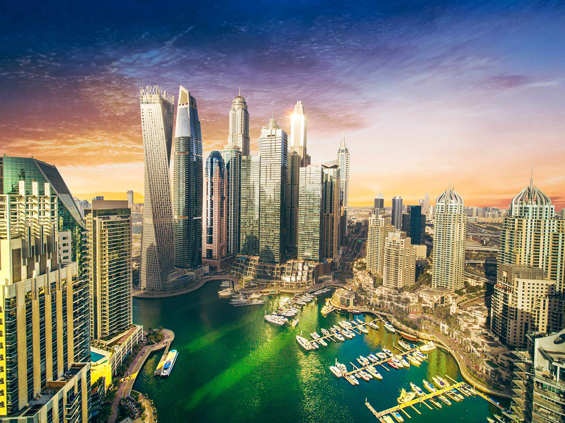 Das Foto zeigt die Dubai Marina bei Sonnenuntergang, mit markanten Wolkenkratzern wie dem Cayan Tower und dem Yachthafen im Vordergrund.